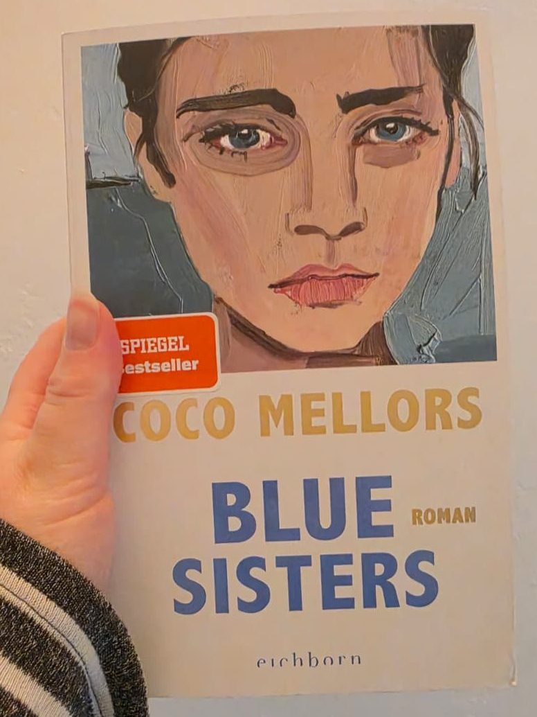 Eine Hand hält den Roman Blue Sisters von Coco Melors. Auf dem Cover ist das Bild einer Frau zu sehen. Es ist ein Spiegel Beststeller Sticker auf dem Cover.