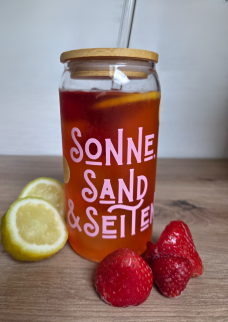 Glas mit bunter Aufschrift, gefüllt mit einem roten Getränk und einem Strohhalm. Um das Glas liegen Erdbeeren und Zitronen.